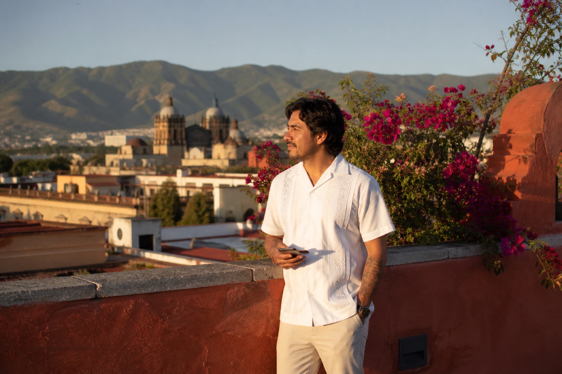 Mexican traveler on a sunlit Oaxaca rooftop with terracotta walls, bougainvillea, and church domes in the distance